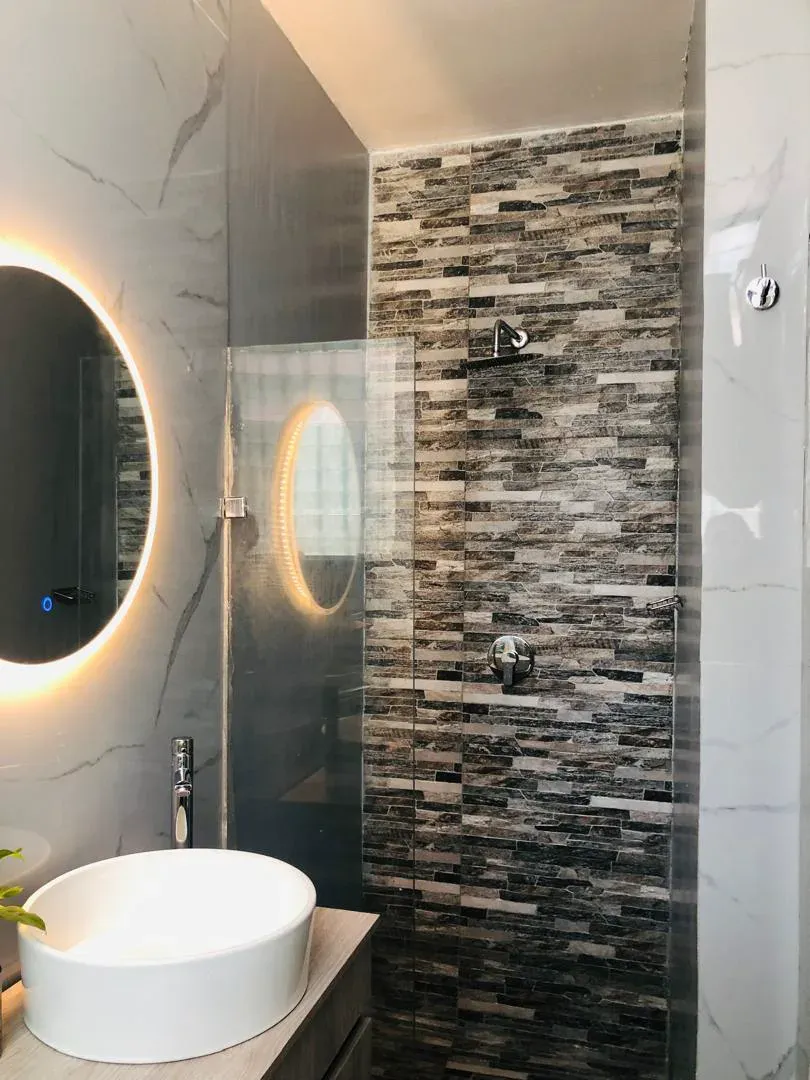Ducha con mampara de vidrio y frente de mosaico gris, junto a lavabo con espejo circular iluminado