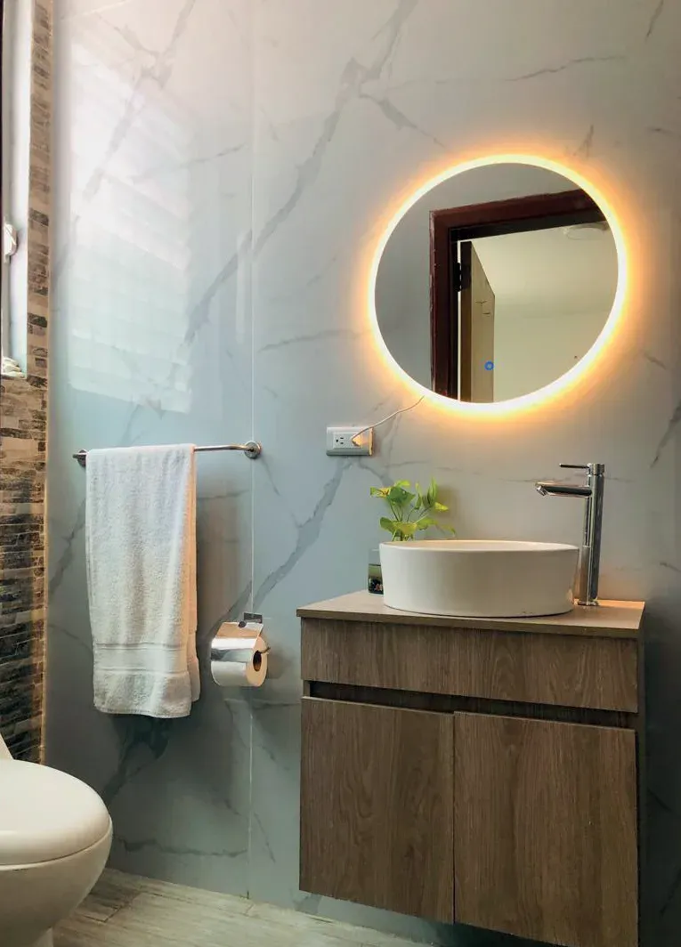 Baño con espejo circular retroiluminado, mueble suspendido de madera y lavabo sobre encimera de mármol blanco